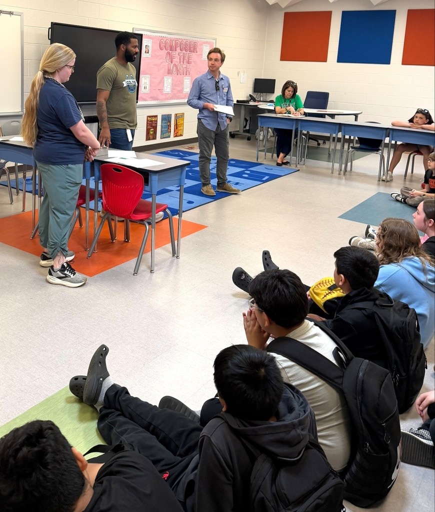Group of students sitting on the floor listening to adults speak.