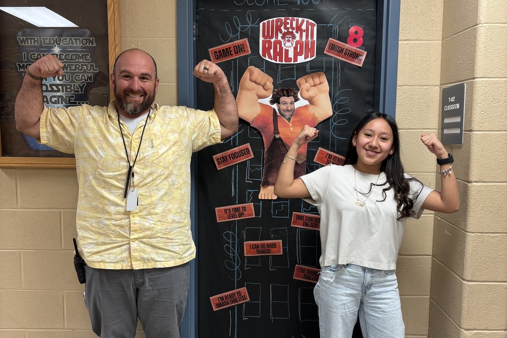 An admin and student are in the hallway flexing their muscles in front of a door with encouraging messages for TCAP.