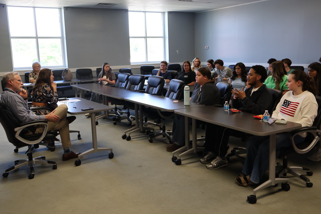 The superintendent and assistant superintendent are talking to the group of students who are seated in chairs. 