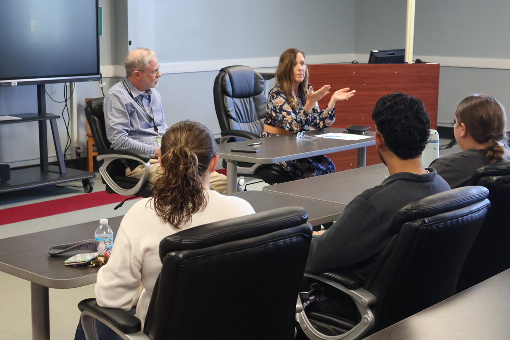 The superintendent and assistant superintendent are talking to the group of students who are seated in chairs. 