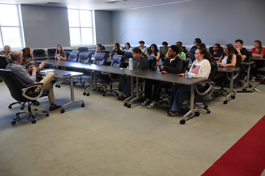 The superintendent and assistant superintendent are talking to the group of students who are seated in chairs. 