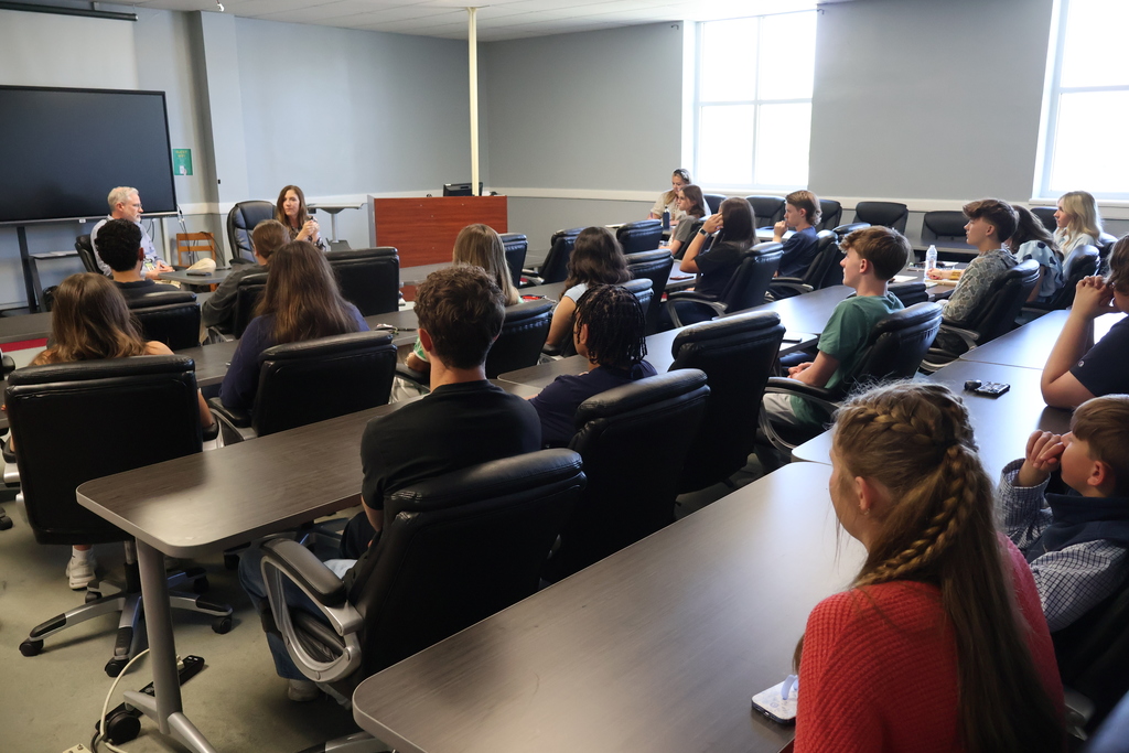 The superintendent and assistant superintendent are talking to the group of students who are seated in chairs. 