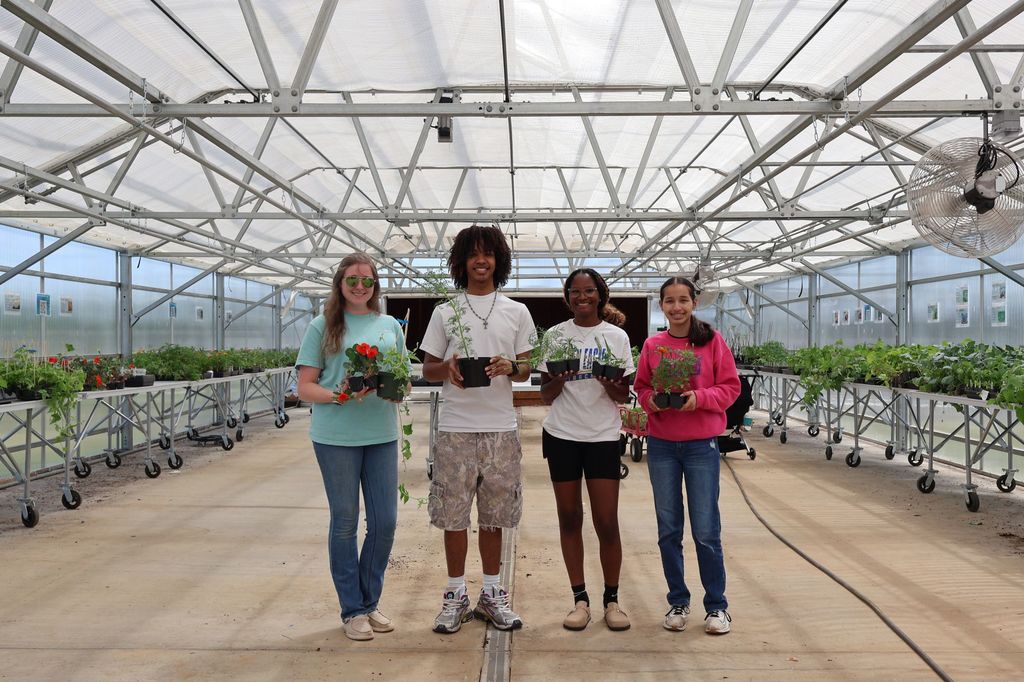 Group of students and their teacher are holding plants in the greenhouse.