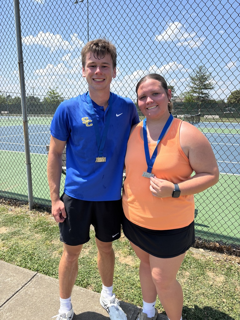 Two student atheletes stand near the tennis court wearing medals.