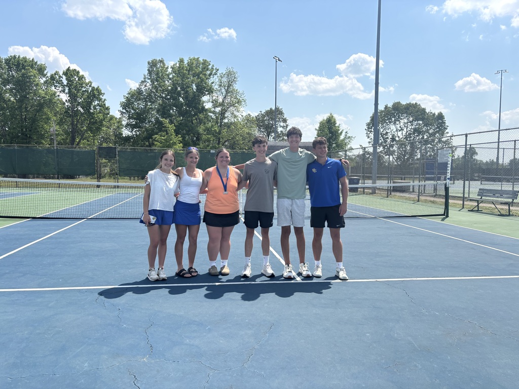 A group of student atheletes are standing on the tennis court for a group picture. 