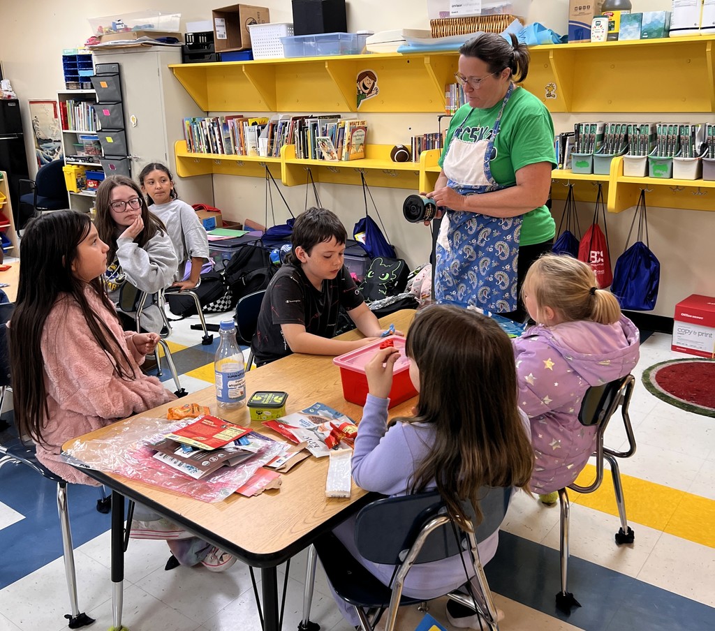 Students are sitting at tables listening to the teacher speak. 