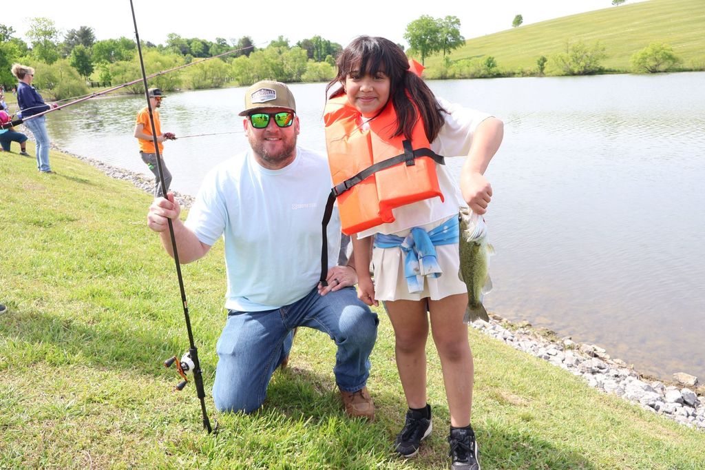 Student is holding a fish and adult helping is holding a fishing pole.