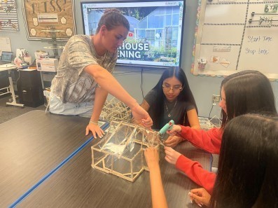 A teacher is showing students how to put a structure together with hotglue and wooden sticks.