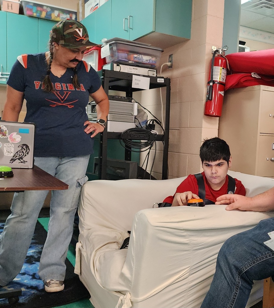 A student is sitting on a couch and pushing an orange button with other two other people nearby. 