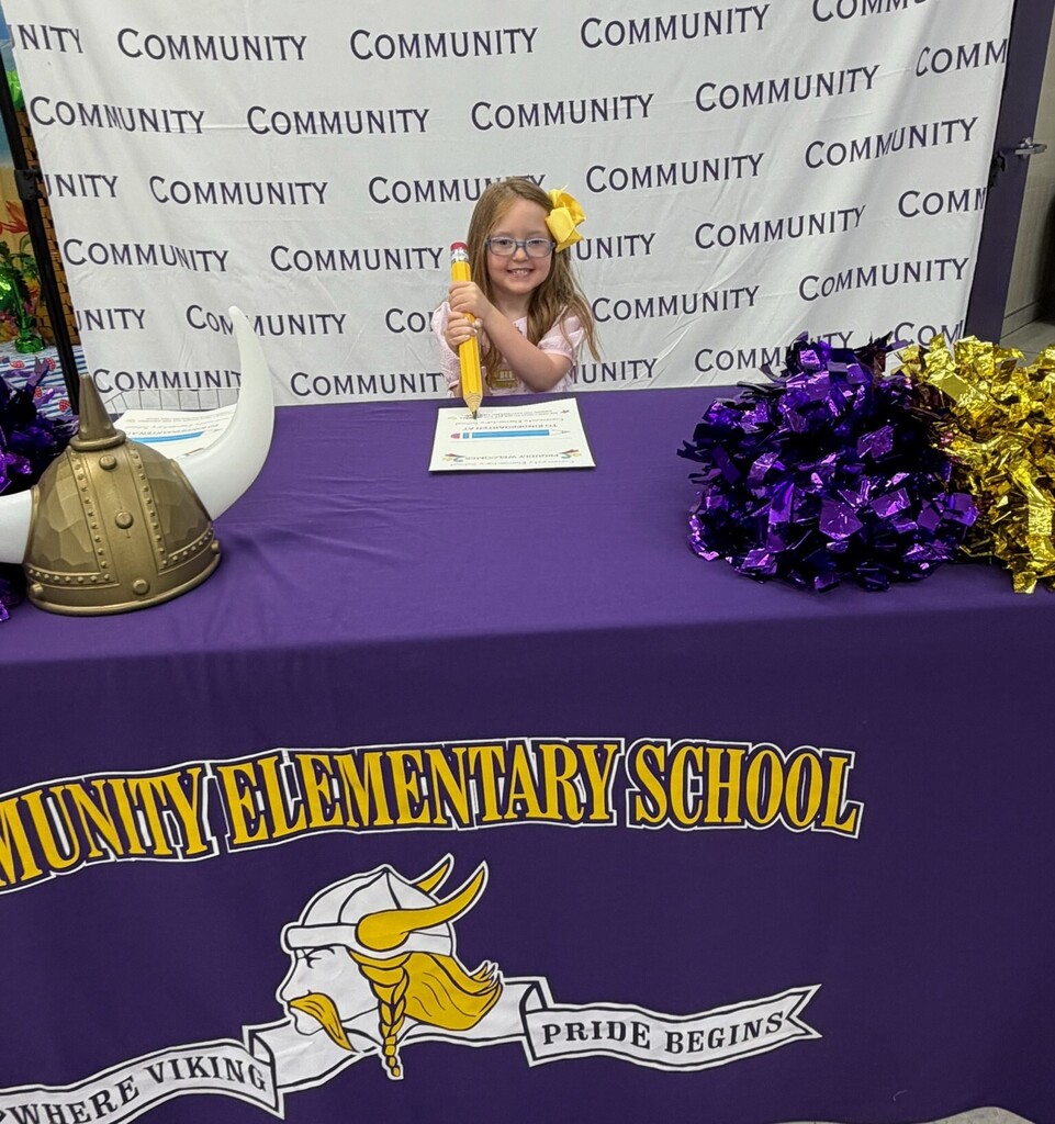 A girl is standing at a table with a big pencil about to write on a piece of paper at Community Elementary School. 