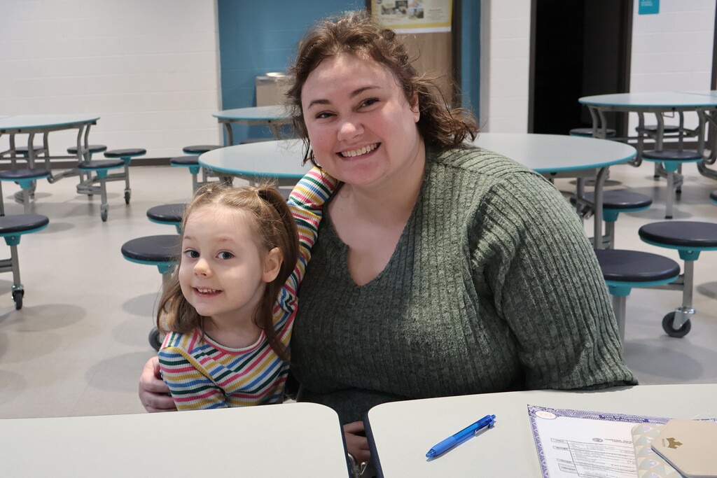 A girl and her mom pose for a picture sitting down at the tables in the cafeteria.