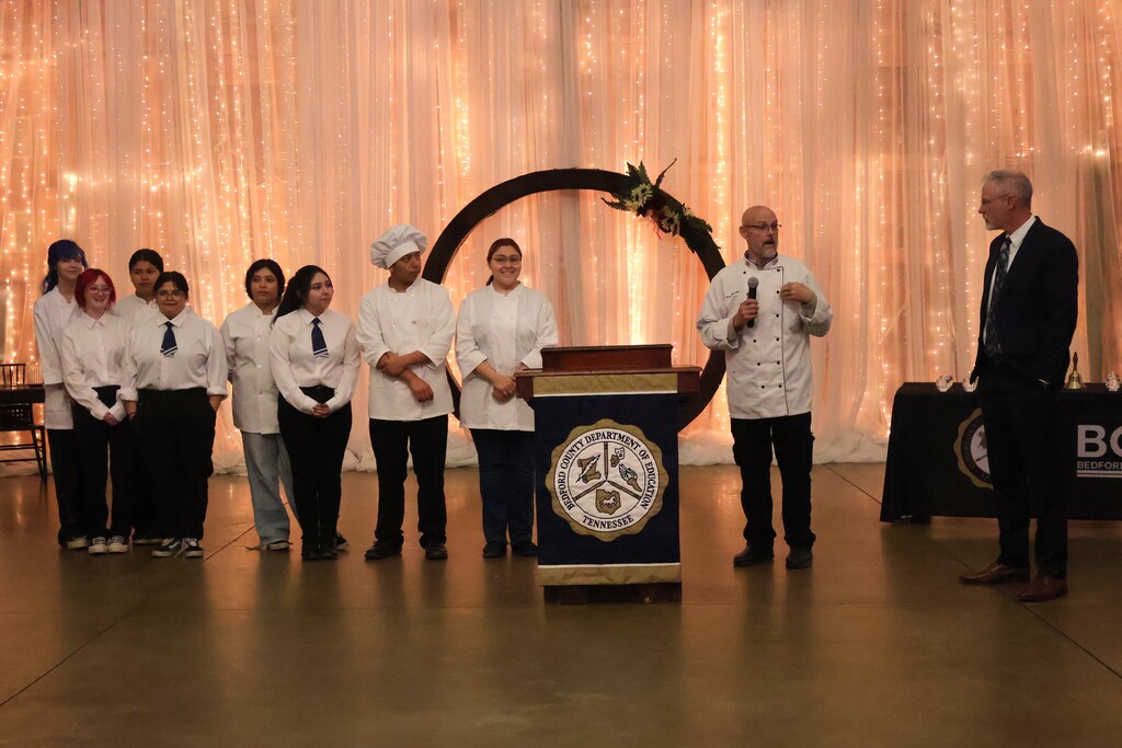 The chef, along with his students, stand with the incoming superintendent so that they can be recognized for cooking and serving the food.