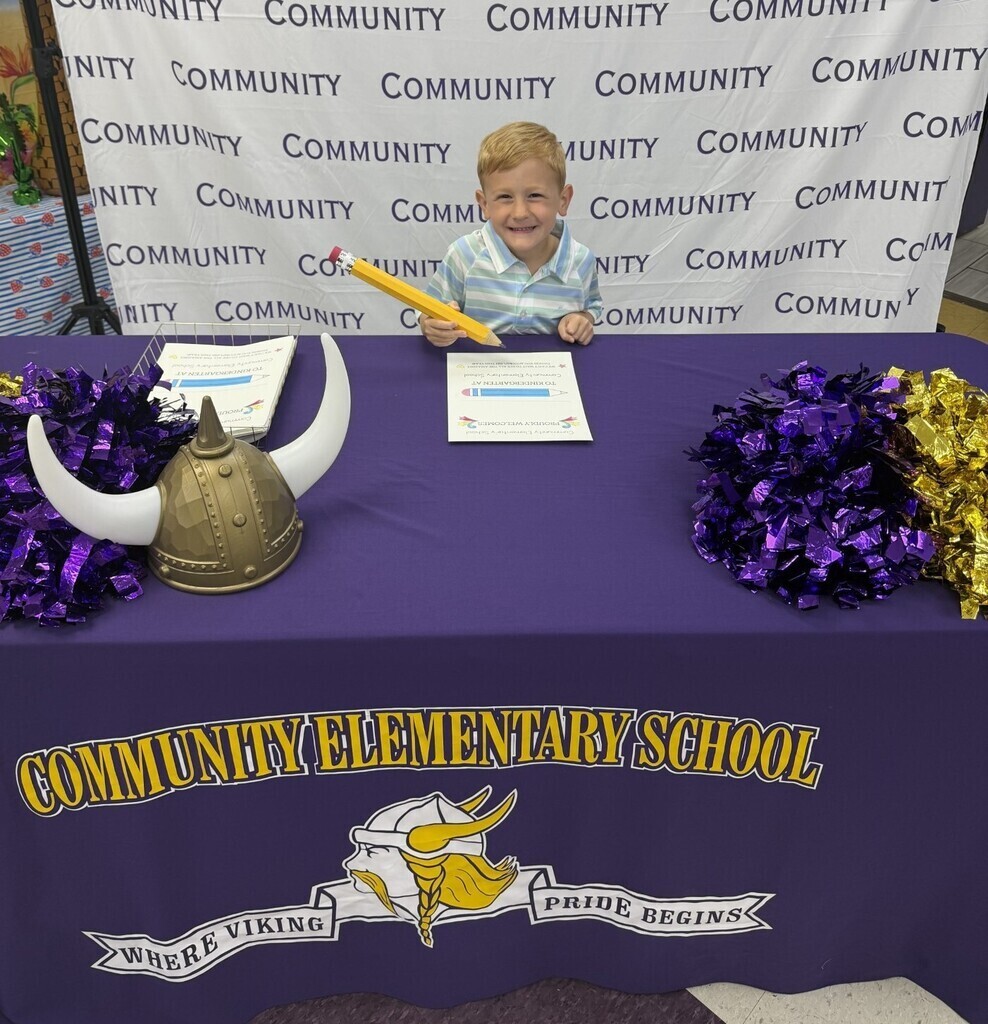 A boy is holding a large pencil while signing a piece of paper at Community Elementary School