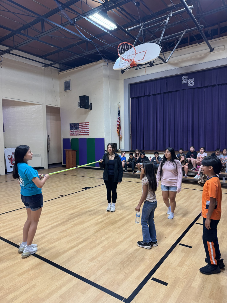 Three elementary students participate in a llimbo activity as two TAP students help hold the pole. 