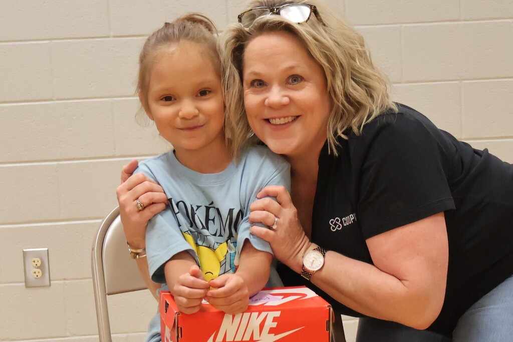 A student, sitting in a chair with a woman from Cooper Steel standing beside her,  is excited about the new shoes she has received.