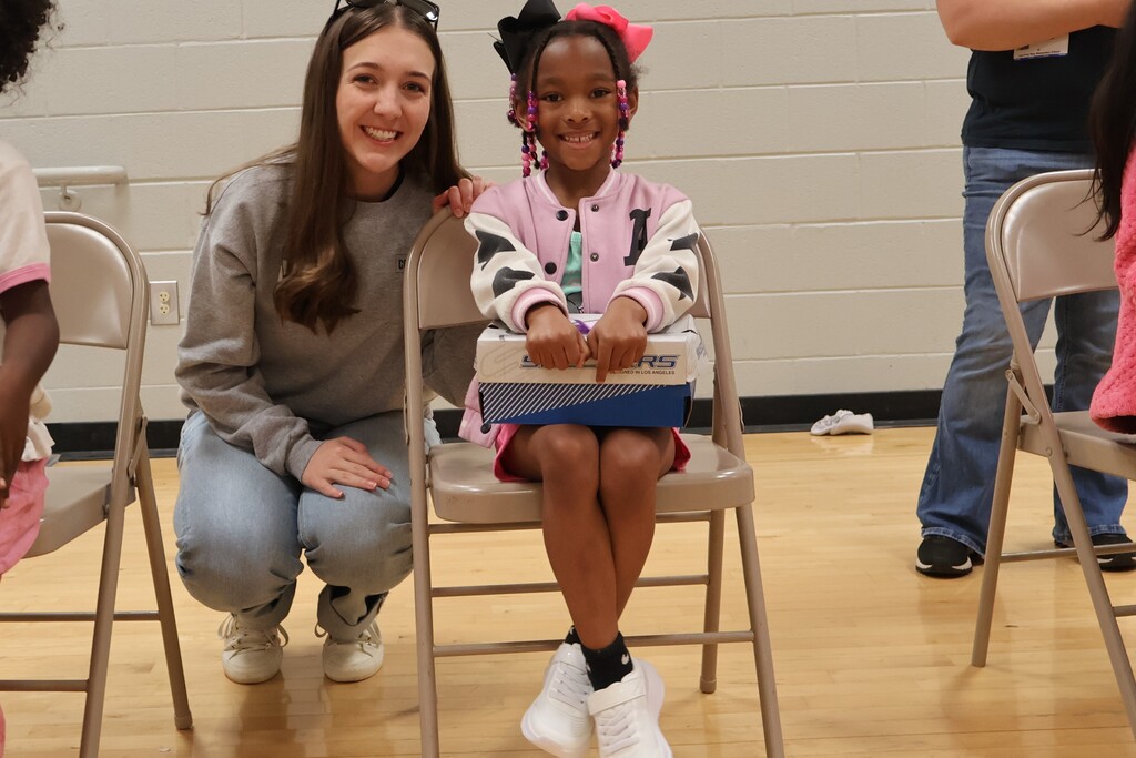 A student is excited over the new shoes she just received as an employee from Cooper Steel is kneeled down beside her.