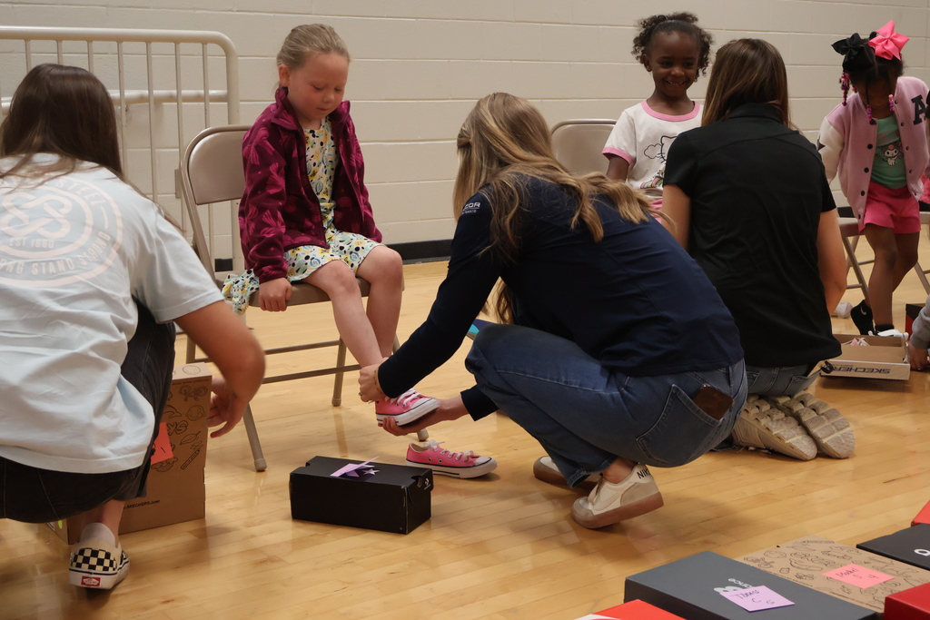 Several students are sitting in chairs trying on their new shoes with the employees from Cooper Steel kneeled down in front of them to assist them. 