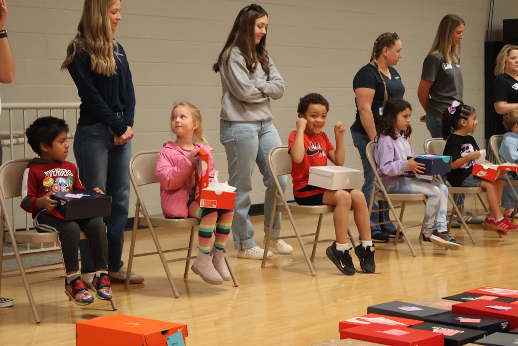 Students are sitting in chair with community partners behind them as they wait to try on their new shoes.
