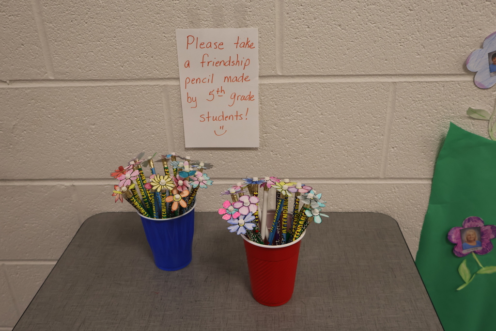 There are two cups sitting on the table with a sign on the wall that says "Please take a friendship pencil made by 5th grade students!". 