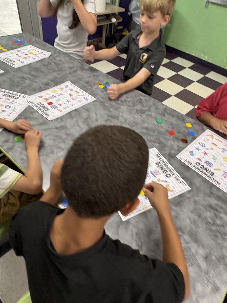 There are several students sitting down at a table with their bingo sheets and the pieces that they will use to mark their sheets while playing bingo.