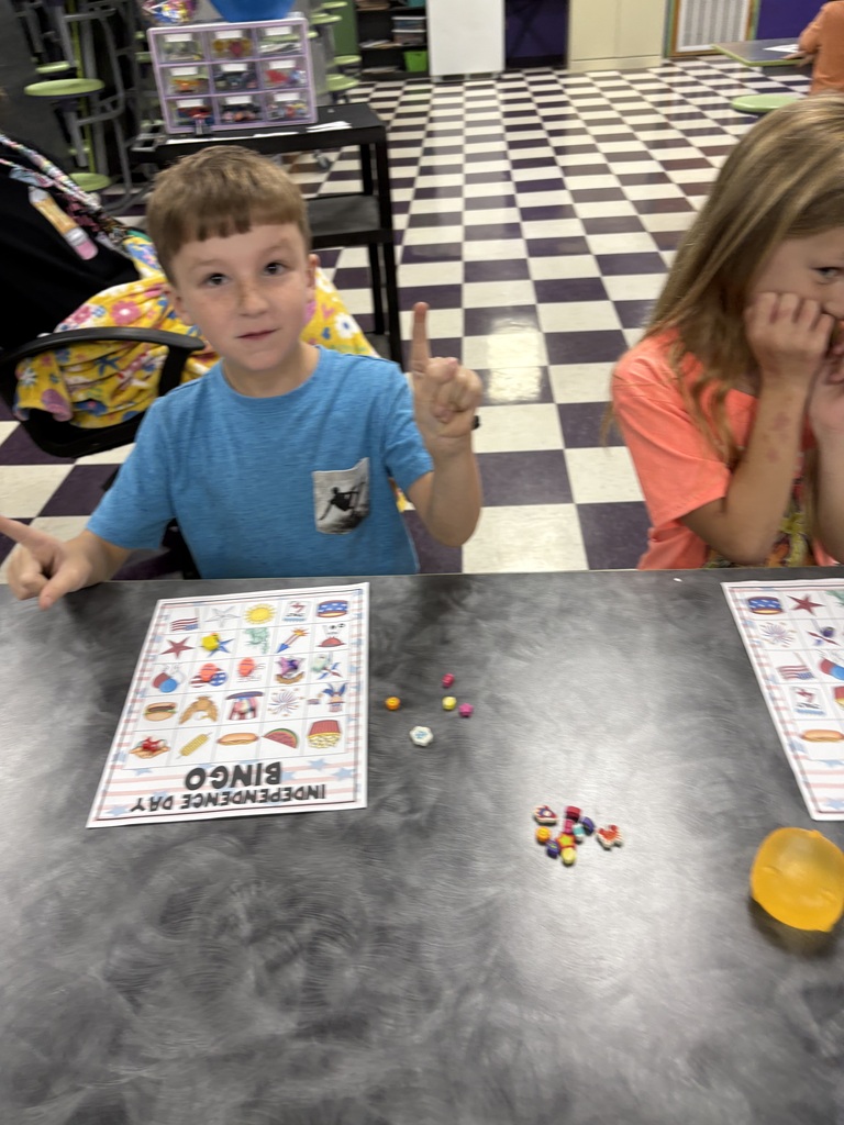 There are two students sitting down at a table with their bingo sheets and the pieces that they will use to mark their sheets.