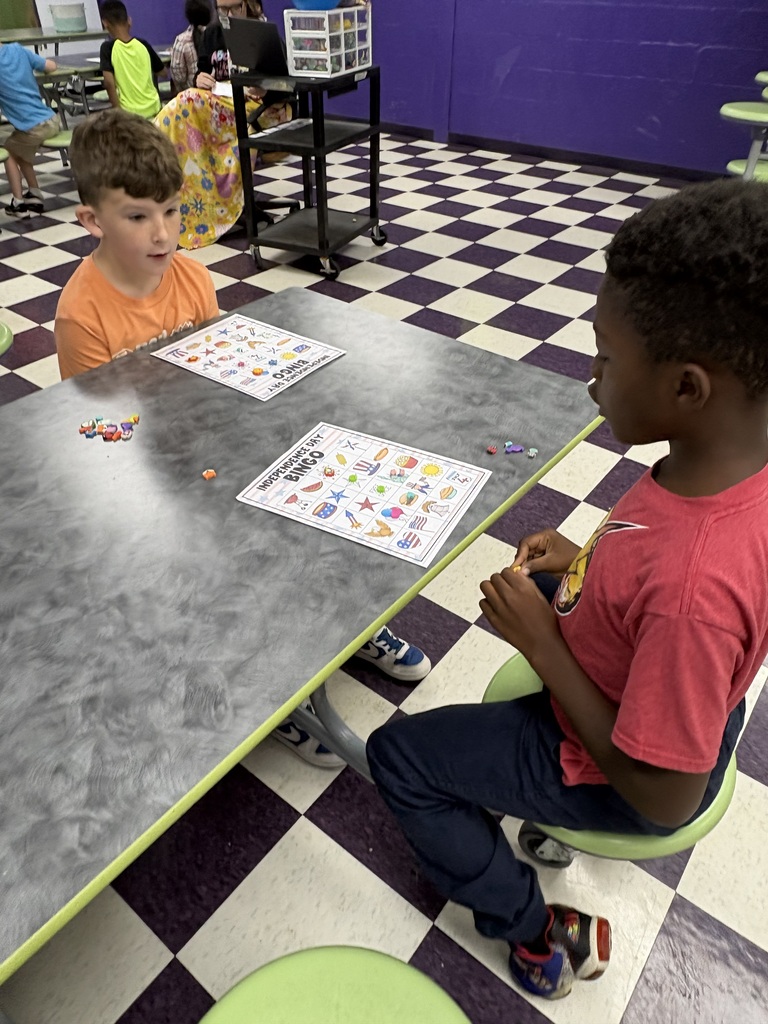 There are two students sitting down at a table with their bingo sheets and the pieces that they will use to mark their sheets.