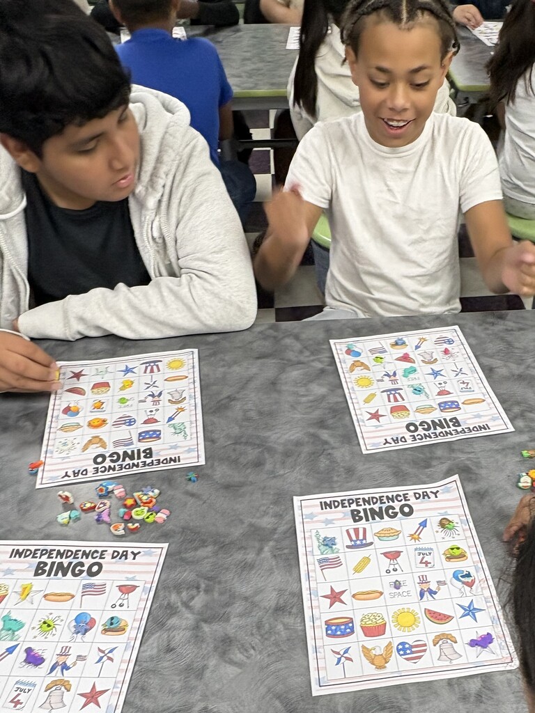 There are two students sitting down at a table with their bingo sheets and the pieces that they will use to mark their sheets.