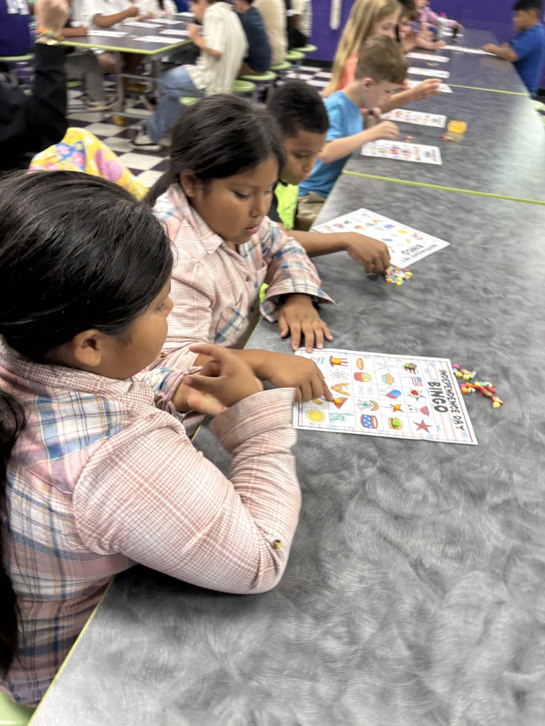 There are several students sitting down at a table with their bingo sheets and the pieces that they will use to mark their sheets while playing bingo.