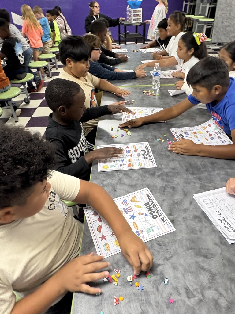There are several students sitting down at a table with their bingo sheets and the pieces that they will use to mark their sheets while playing bingo.