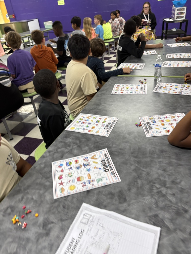 There are several students sitting down at a table with their bingo sheets and the pieces that they will use to mark their sheets while playing bingo.