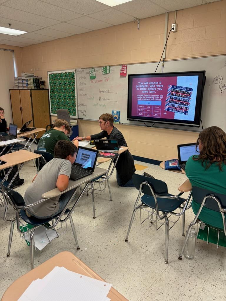 Students are sitting at their desks while the teacher is explaining something to a student as she kneels on the ground.