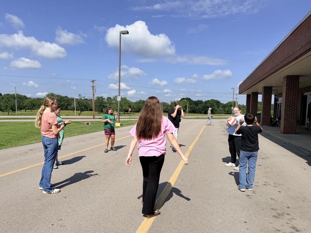 There are seven students playing volleyball together out by the school.