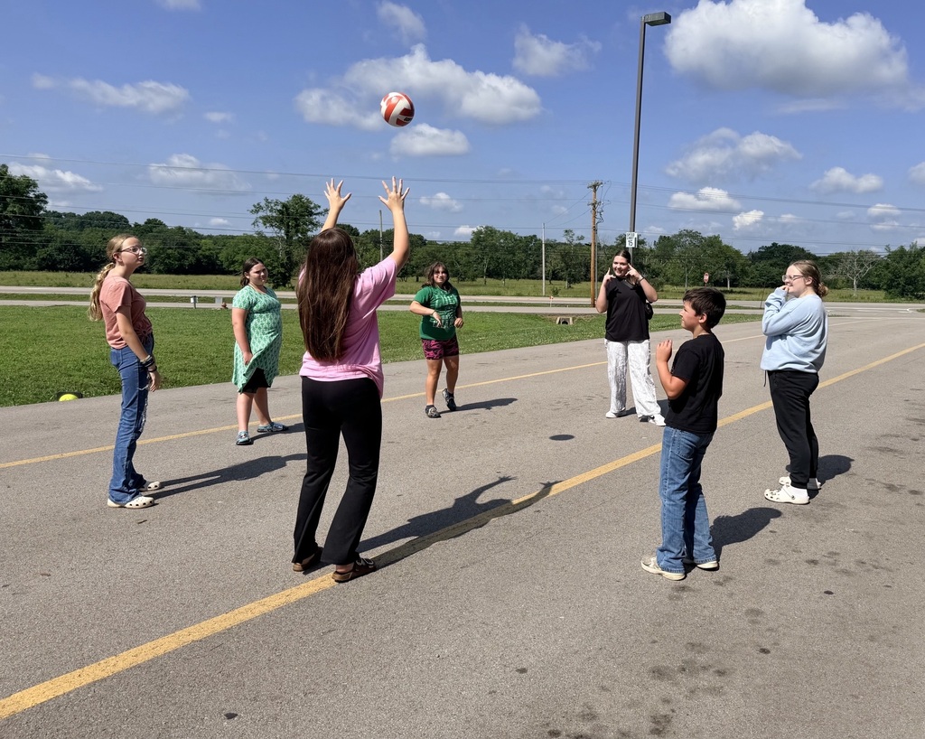 There are seven students playing volleyball together out by the school.