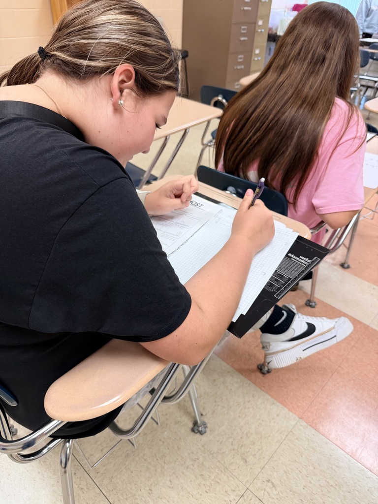 There are two students seated at their desks while doing work on paper.