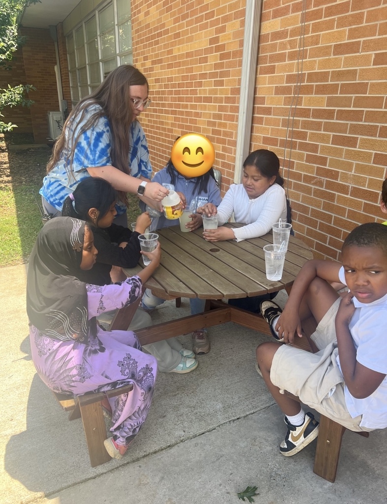 There are several students, along with their teacher, sitting in chairs in the shade enjoying the lemonade that they made.