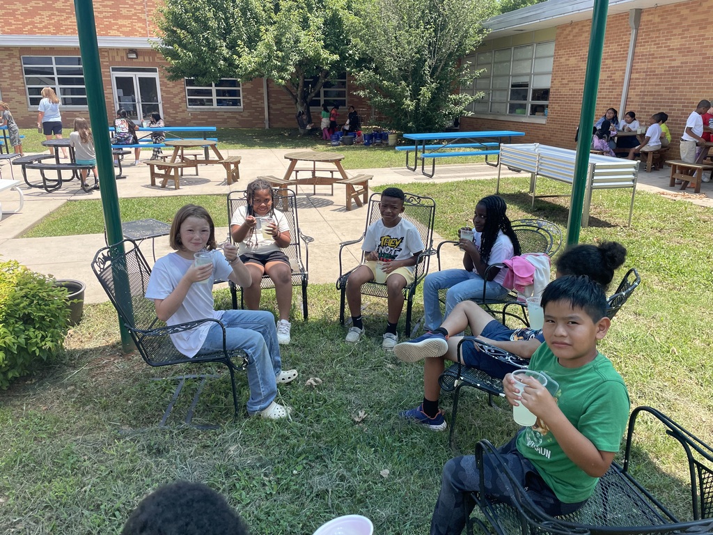 There are several students sitting in chairs in the shade enjoying the lemonade that they made.