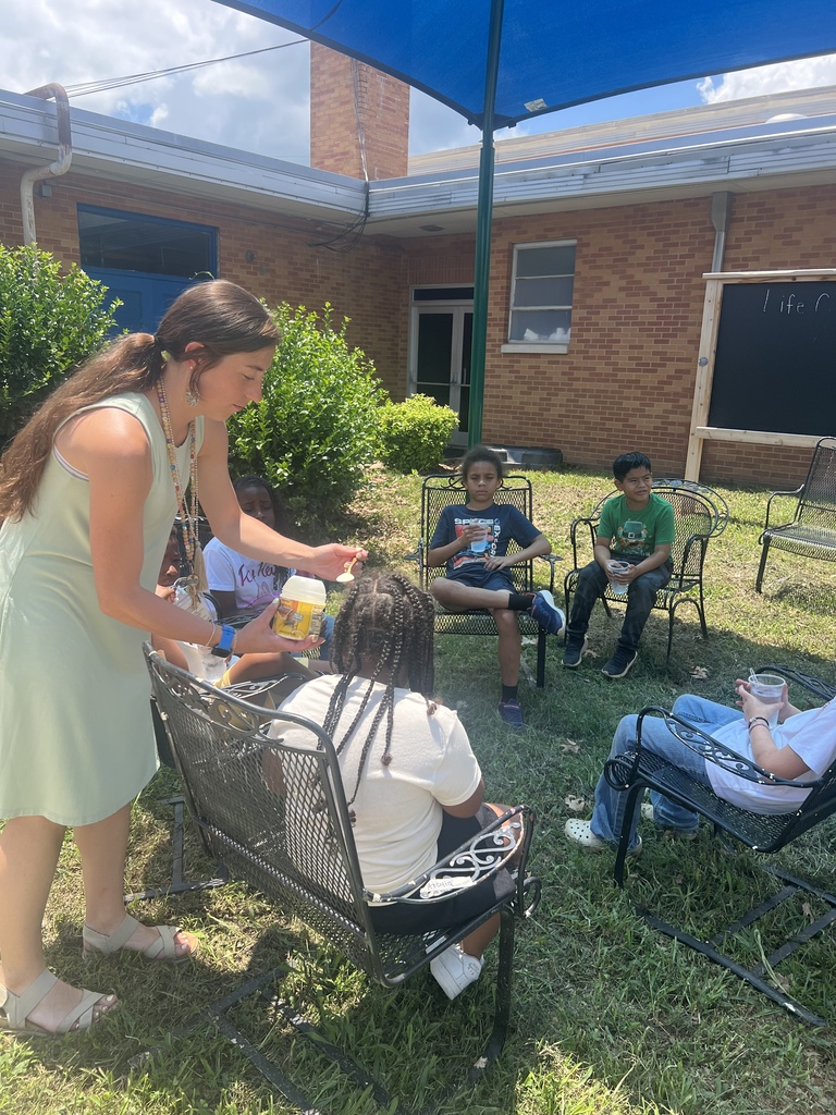 There are several students, along with their teacher, sitting in chairs in the shade enjoying the lemonade that they made. 