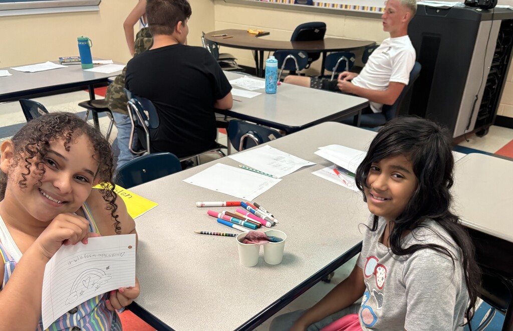 Two students are sitting down at a table with two cups of colored water as they participate in the walking water experiment. 