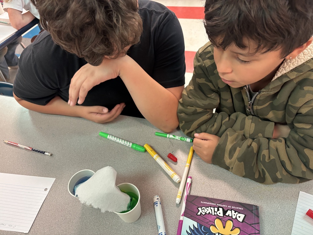 Two students are sitting down at a table with two cups of colored water as they participate in the walking water experiment. 