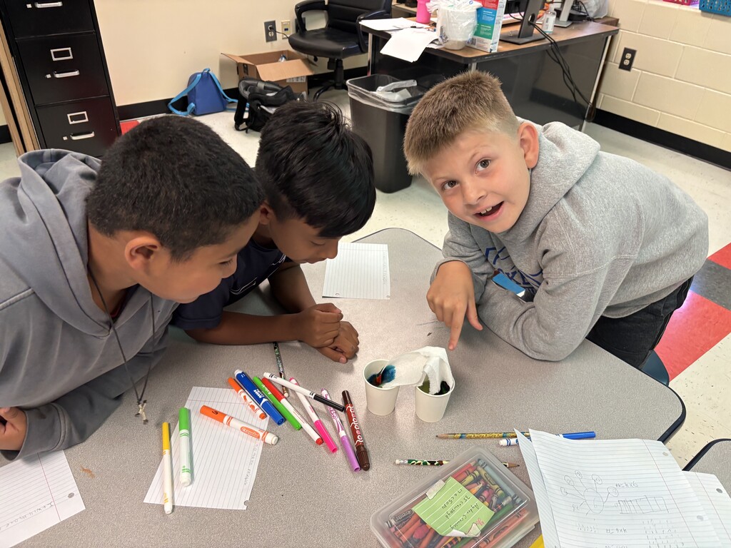 Three students are sitting down at a table with two cups of colored water as they participate in the walking water experiment. 
