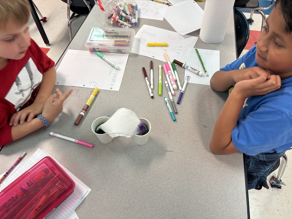 Two students are sitting down at a table with two cups of colored water as they participate in the walking water experiment. 