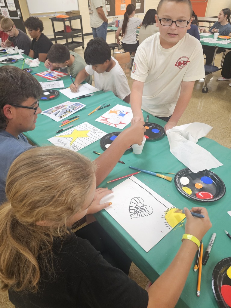 Several students are sitting at a table painting their drawings that they made to celebrate America's 250th anniversary. 