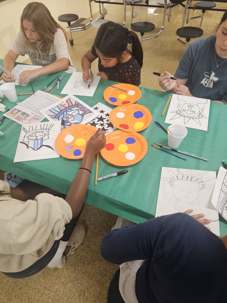 Several students are sitting at a table painting their drawings that they made to celebrate America's 250th anniversary. 