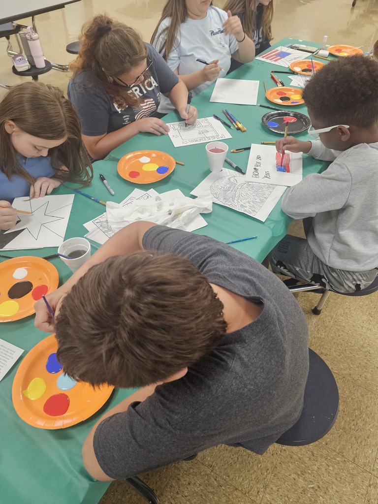 Several students are sitting at a table painting their drawings that they made to celebrate America's 250th anniversary. 