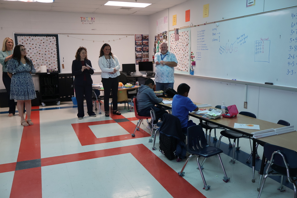 Students are sitting at their desks as staff members  and their teacher looks on.