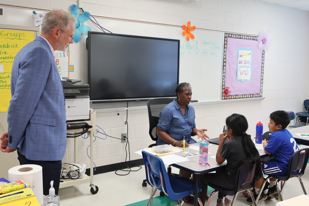 A teacher is sitting at a table with two students teaching as Superintendent Tim Harwell looks on. 