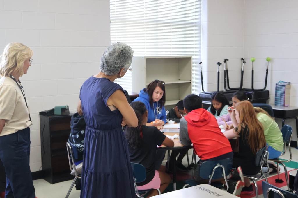 Students are sitting at a large table with their teacher as the Commissioner looks on. 