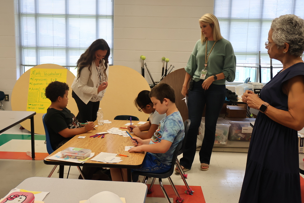 Three students are sitting at their desks working as the Commissioner and others look on. 
