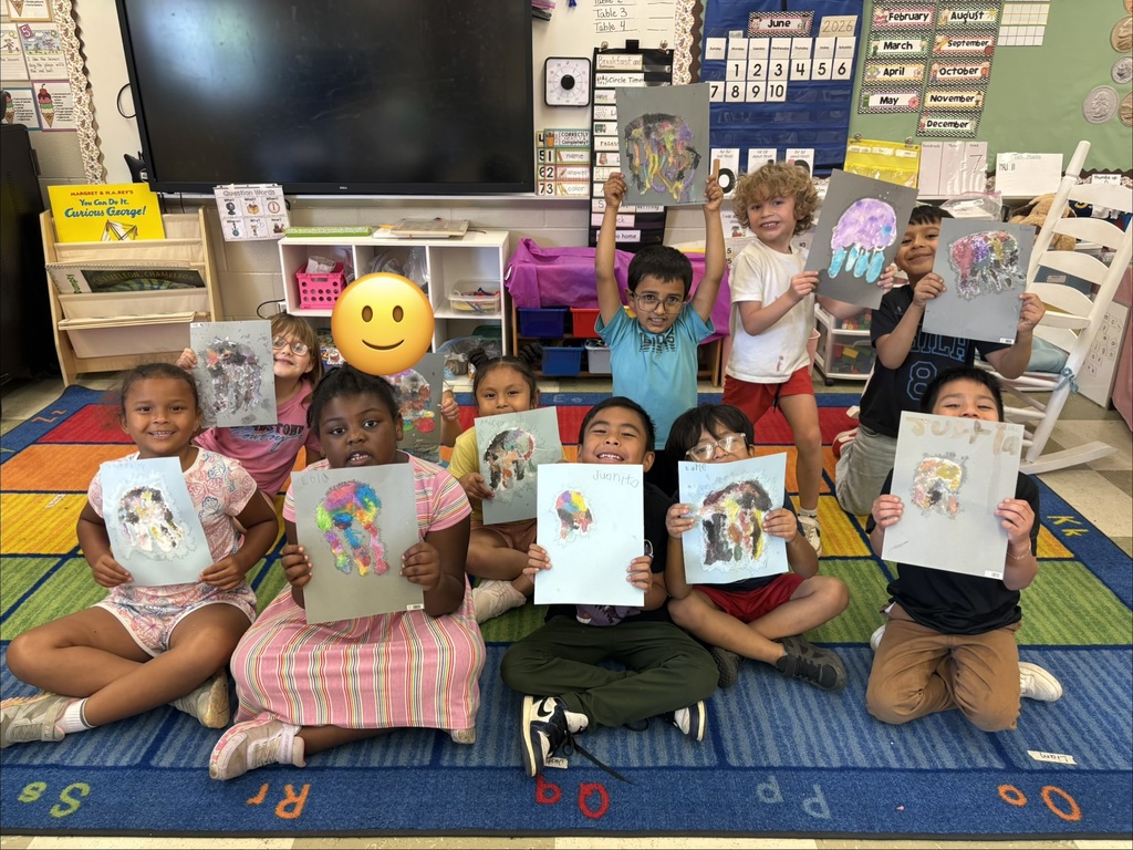 Students are posed for a group picture on the carpet in their classroom holding up the pictures that they created. 