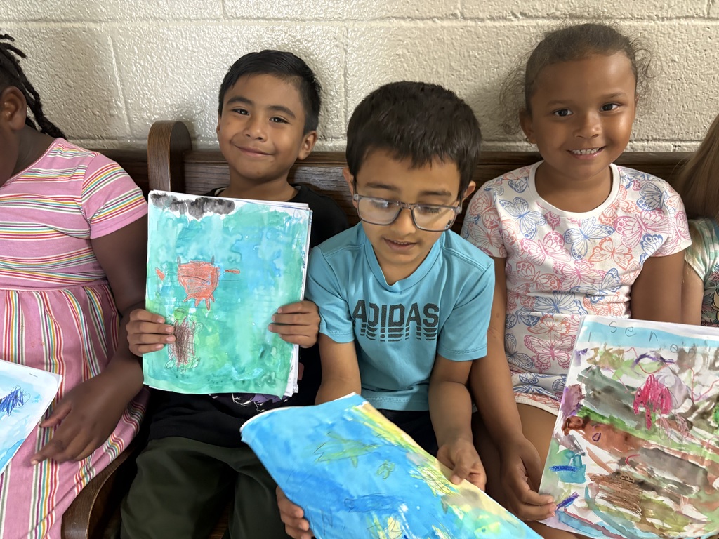 Students are seated on a bench holding up the pictures they created. 